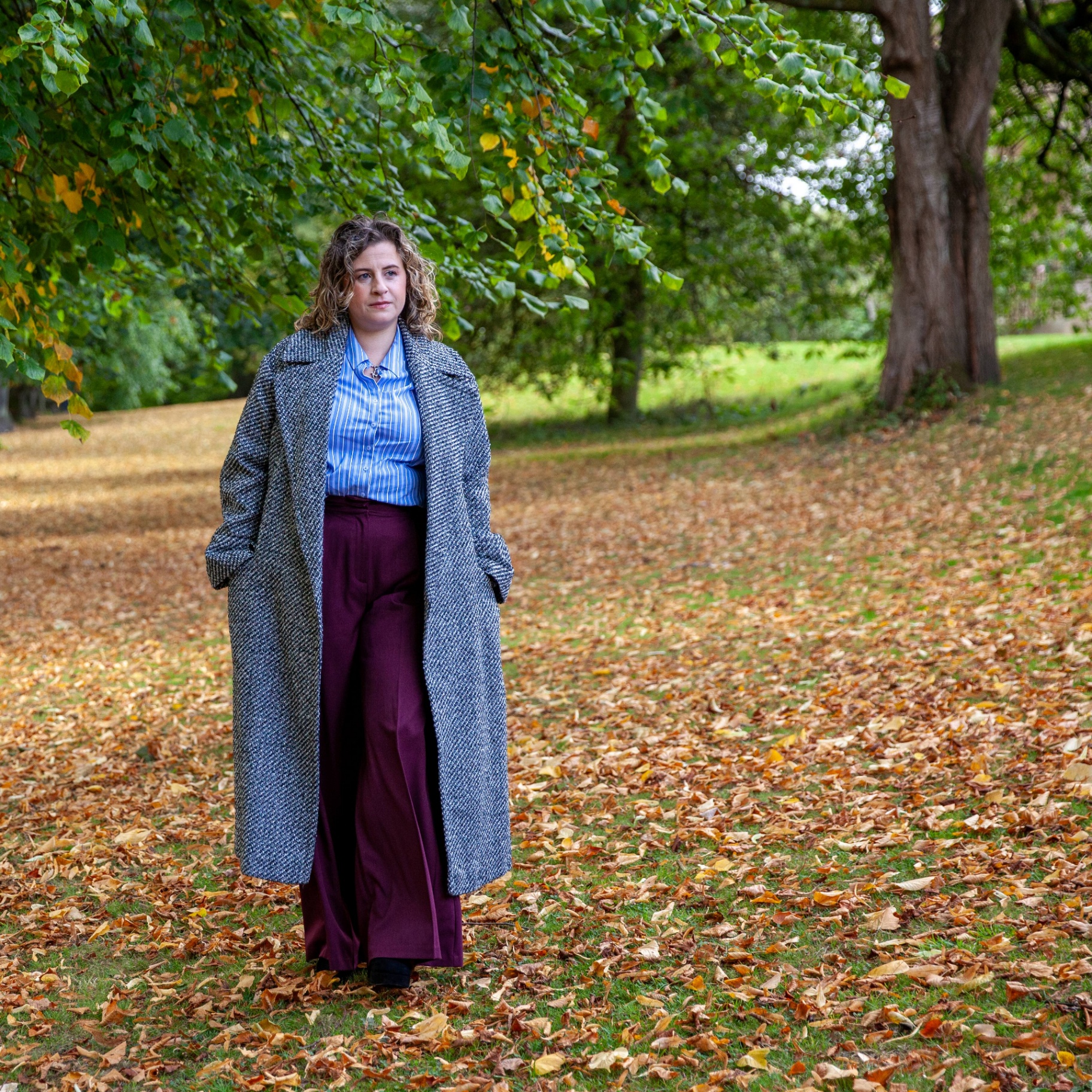 Kate Keir standing outdoors in a long coat among autumn leaves.