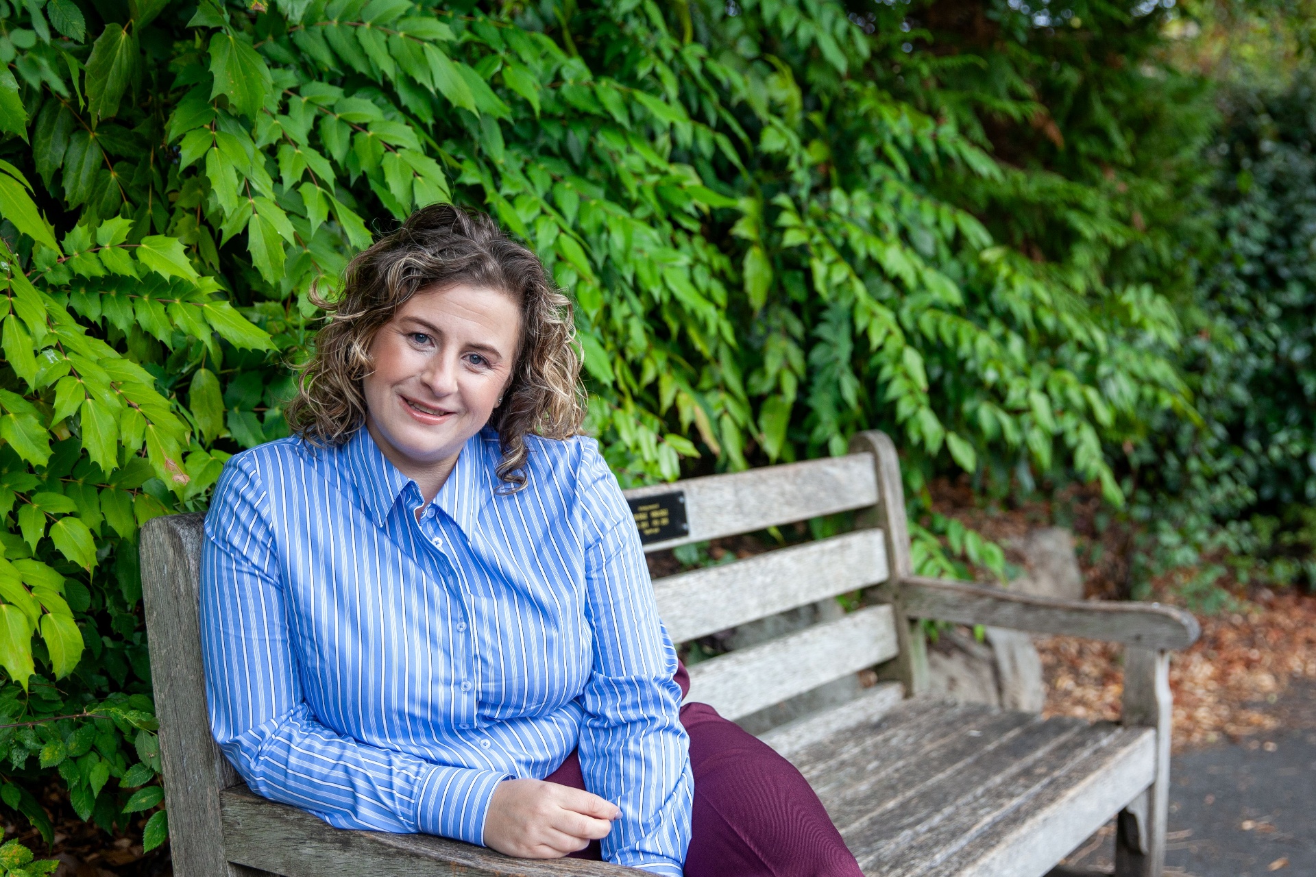 Kate Keir sitting on a wooden bench outdoors, surrounded by green foliage.